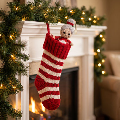 A red and cream striped knit Christmas stocking with a handmade crochet baby safe Mrs. Claus ornament by YarnYummies hanging from a white fireplace mantel decorated with green garland and warm string lights.