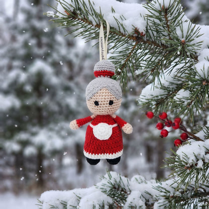 Handmade crochet baby safe Mrs. Claus ornament by YarnYummies, hanging from a snow-covered evergreen branch in a winter outdoor setting.