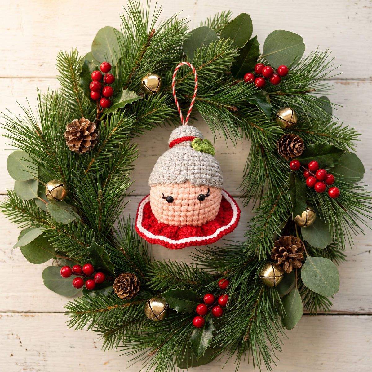 A festive Christmas wreath made of green pine branches and eucalyptus leaves, decorated with red berries, pinecones, small gold bells, and a Handmade crochet Mrs. Claus head ornament with rattle by YarnYummies in the center, set against a white wooden background.