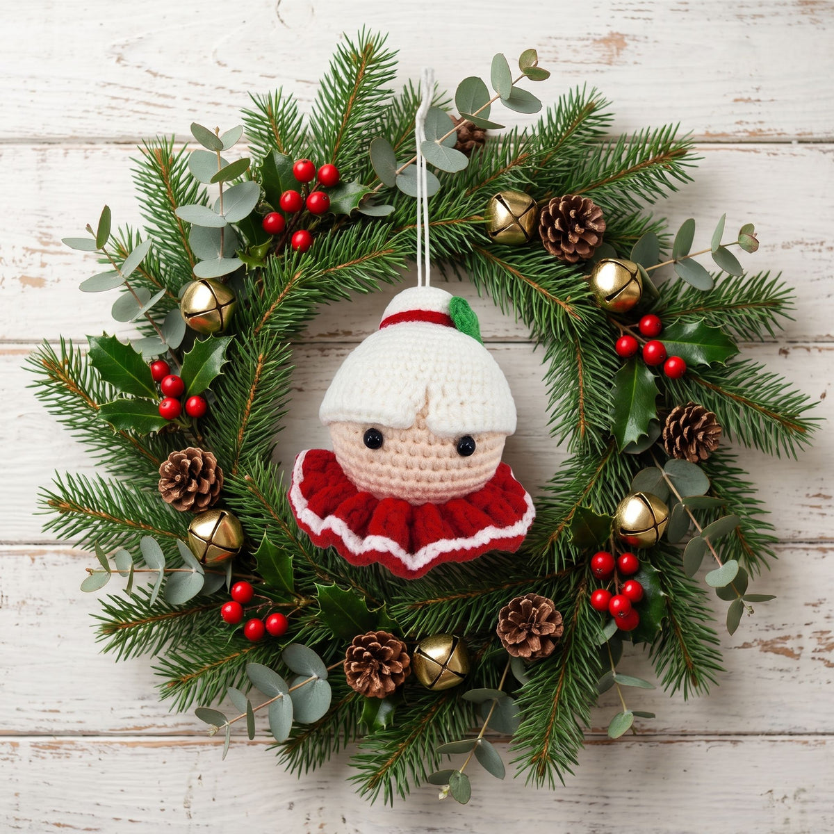 A festive Christmas wreath hanging on a whitewashed wooden wall. The wreath is made of green pine branches with eucalyptus leaves, red berries, pinecones, and small gold bells, and features a central Handmade crochet Mrs. Claus head ornament by YarnYummies front view with a white hat and red ruffled dress.