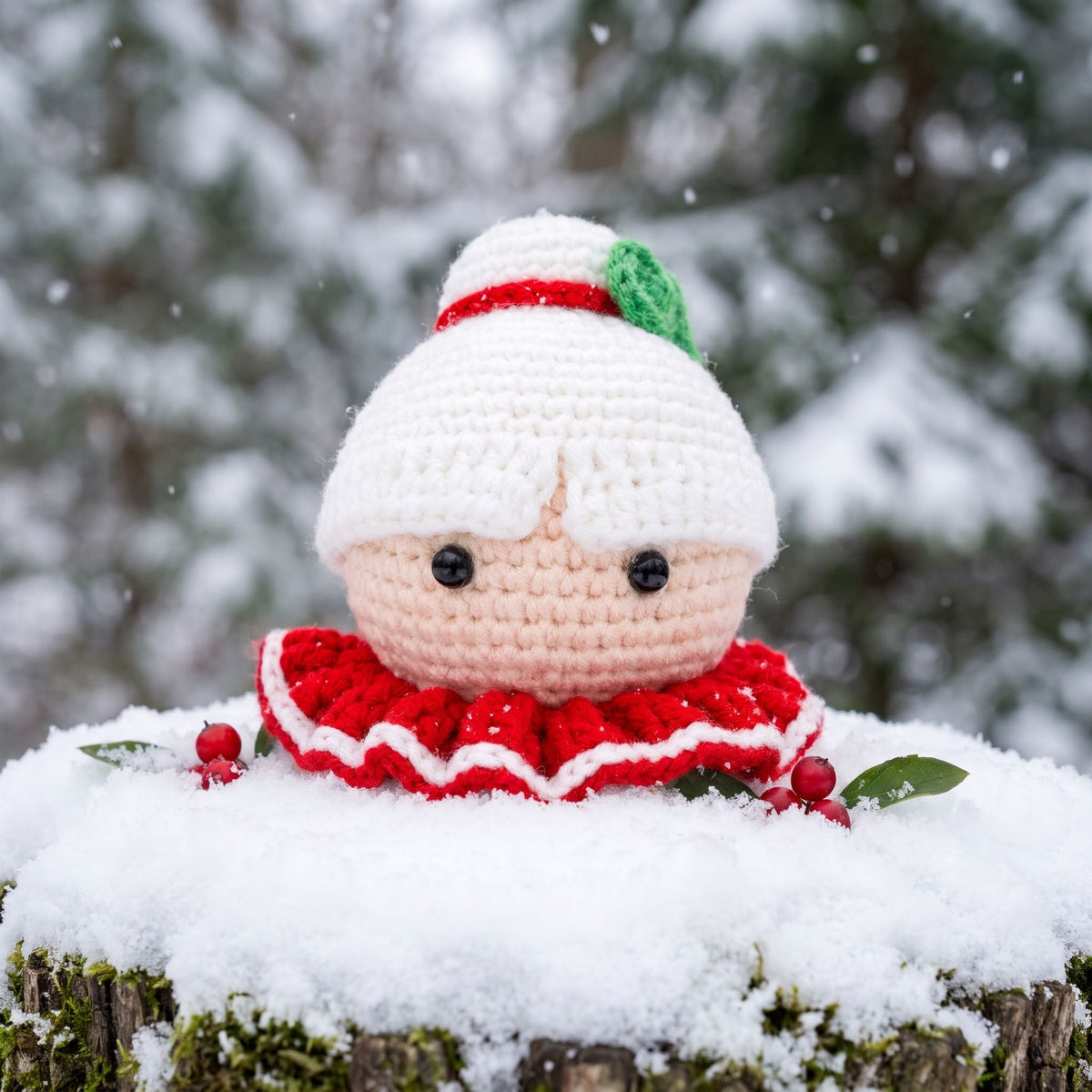 A Handmade crochet Mrs. Claus head ornament by YarnYummies front view with a white hat, red ruffled dress, and peach face sits on a snowy log outdoors with blurred evergreen trees in the background.