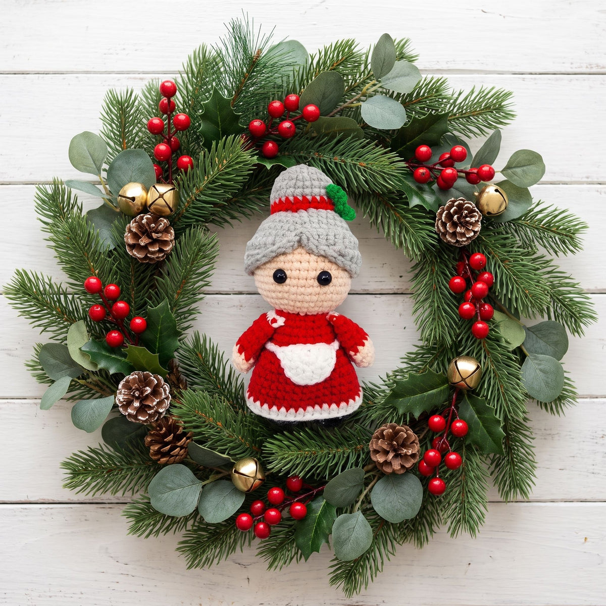 A festive Christmas wreath made of green pine branches with eucalyptus leaves, red berries, pinecones, and gold bells, featuring a Handmade crochet full body Mrs. Claus ornament by YarnYummies in the center, set against a white wood plank background.