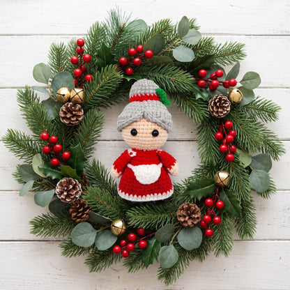 A festive Christmas wreath made of green pine branches with eucalyptus leaves, red berries, pinecones, and gold bells, featuring a Handmade crochet full body Mrs. Claus ornament by YarnYummies in the center, set against a white wood plank background.