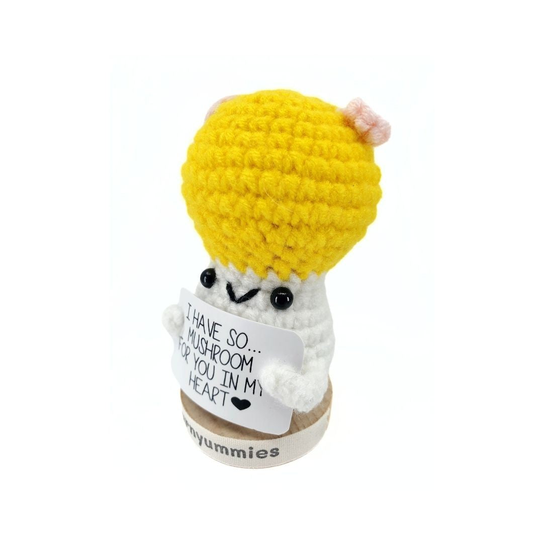 Crocheted mushroom character plush  with a yellow cap and white body, holding a sign.