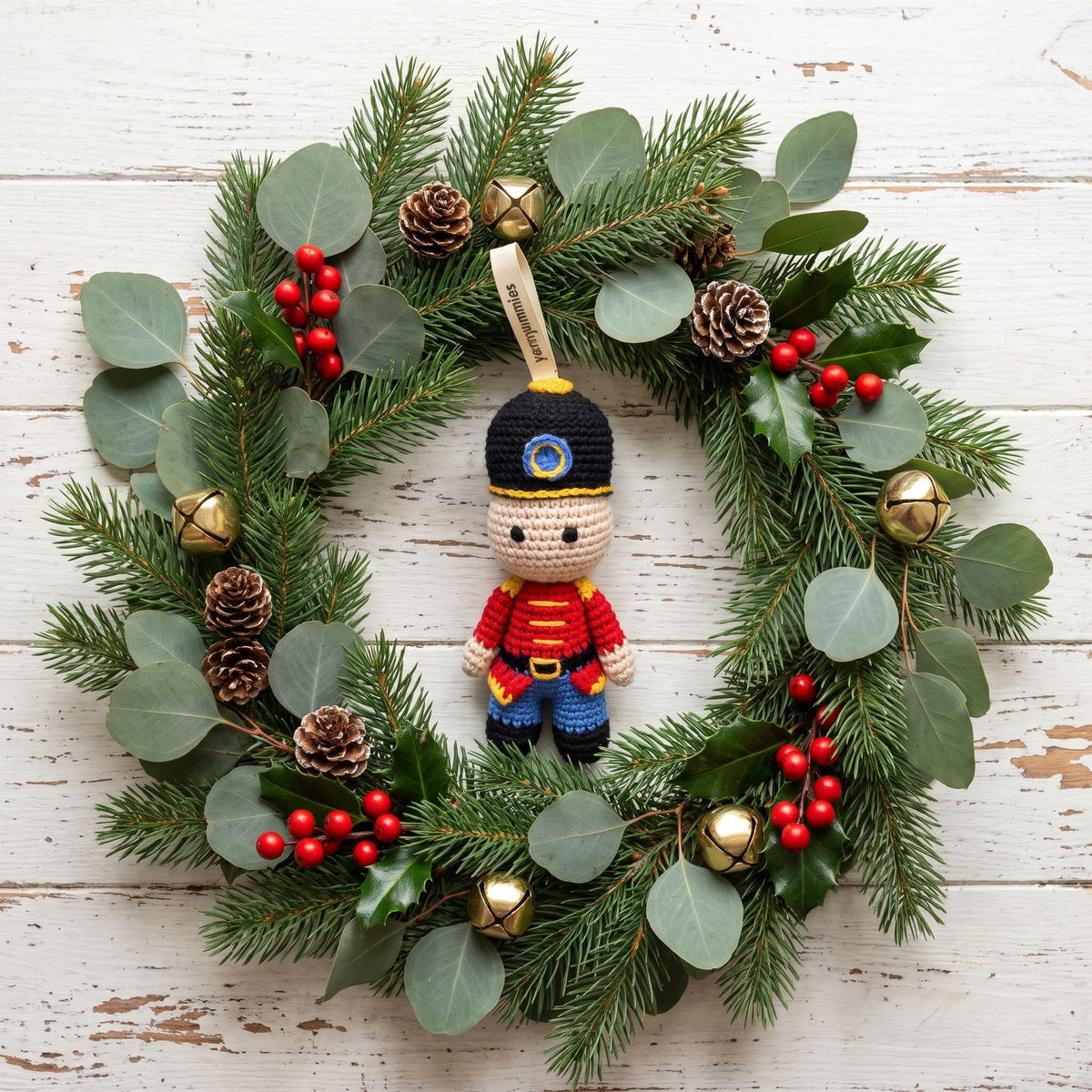A festive Christmas wreath made of green pine branches with eucalyptus leaves, red berries, pinecones, gold bells, and a Handmade crochet baby safe nutcracker ornament by YarnYummies front view showing classic uniform detail in the center, displayed on a white rustic wooden background.