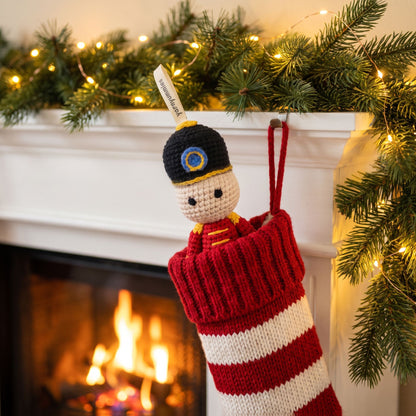 A knitted Christmas stocking hanging from a white fireplace mantel decorated with green pine branches and warm string lights. The stocking is red and white striped with a Handmade crochet baby safe nutcracker ornament by YarnYummies front view showing classic uniform detail peeking out, wearing a black hat with a gold emblem and a red coat.