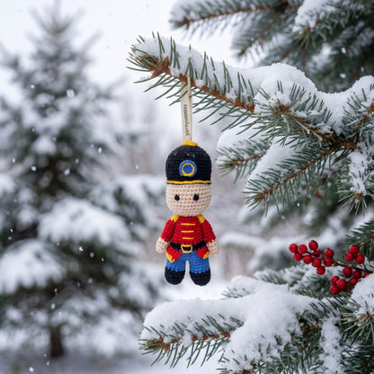 A Handmade crochet baby safe nutcracker ornament by YarnYummies front view showing classic uniform detail hanging from a snow-covered evergreen tree branch in the winter outdoors.