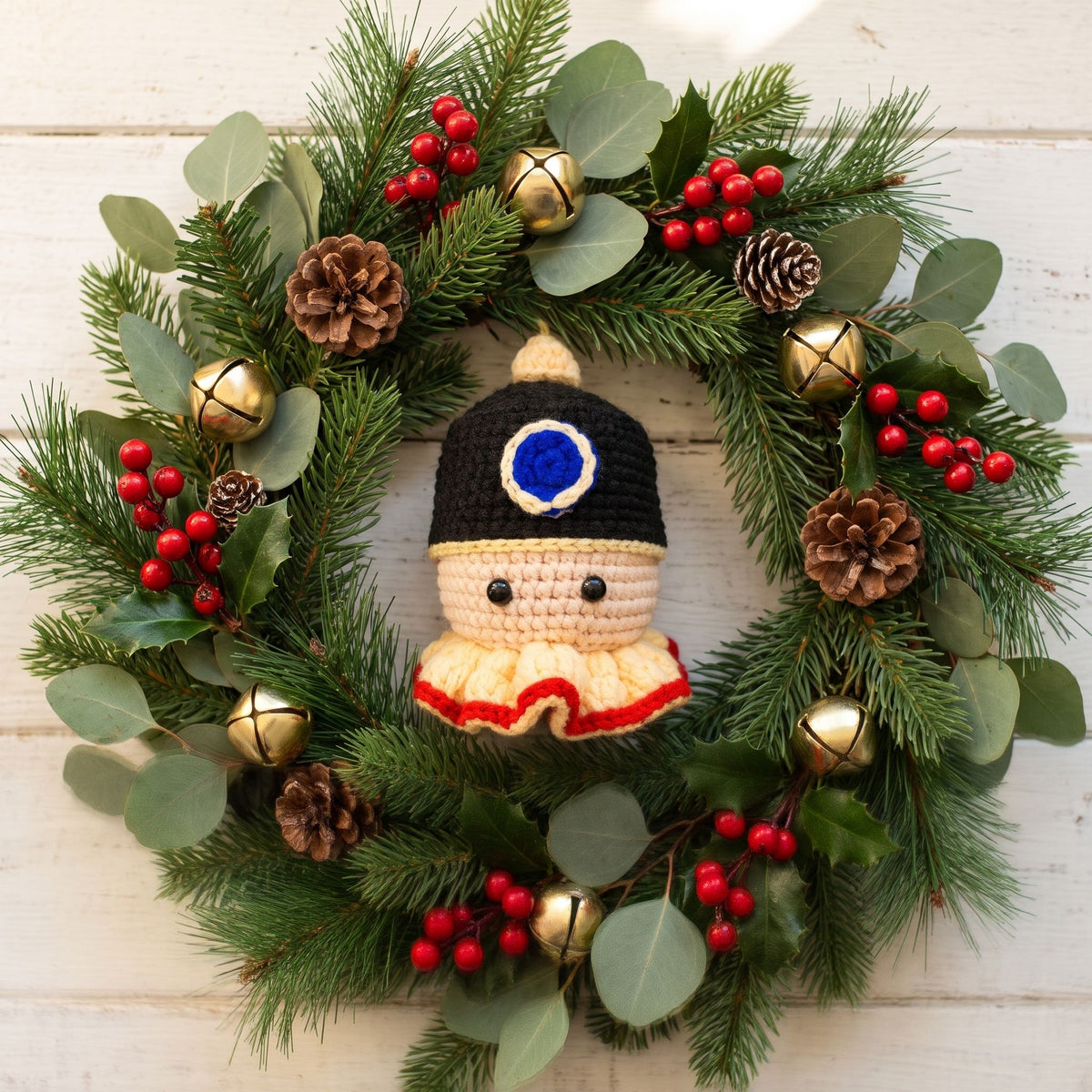 A festive Handmade crochet nutcracker head ornament by YarnYummies front view showing classic hat and face detail centered inside a green pine wreath decorated with red berries, pinecones, eucalyptus leaves, and small gold bells on a white wood plank background.