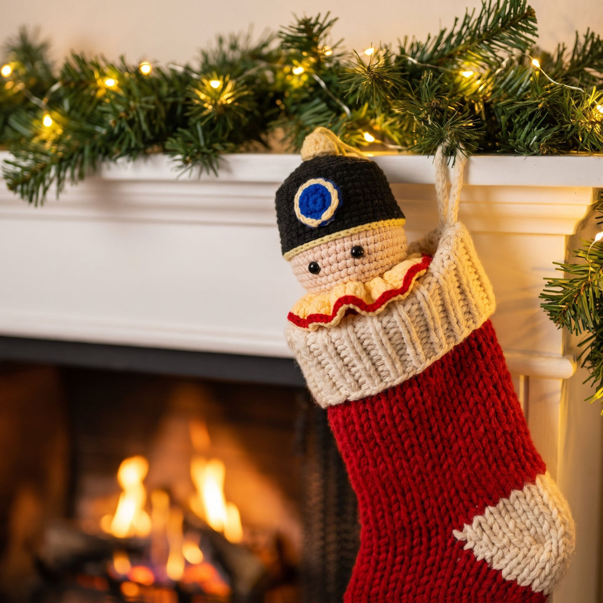 A knitted Christmas stocking hanging from a white fireplace mantel, decorated with a Handmade crochet nutcracker head ornament by YarnYummies front view showing classic hat and face detail peeking out, with a warm fireplace and green garland in the background.