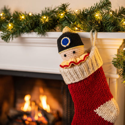 A knitted Christmas stocking hanging from a white fireplace mantel, decorated with a Handmade crochet nutcracker head ornament by YarnYummies front view showing classic hat and face detail peeking out, with a warm fireplace and green garland in the background.