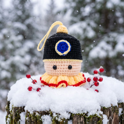 A Handmade crochet nutcracker head ornament by YarnYummies front view showing classic detail in a black and yellow hat with a blue emblem, posed on a snow-covered tree stump outdoors in the snow.