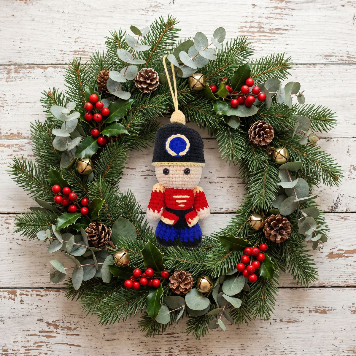 A Handmade crochet full body nutcracker ornament by YarnYummies front view showing uniform detail hanging from a green pine wreath decorated with eucalyptus leaves, pinecones, and red berries, set against a whitewashed wooden background.