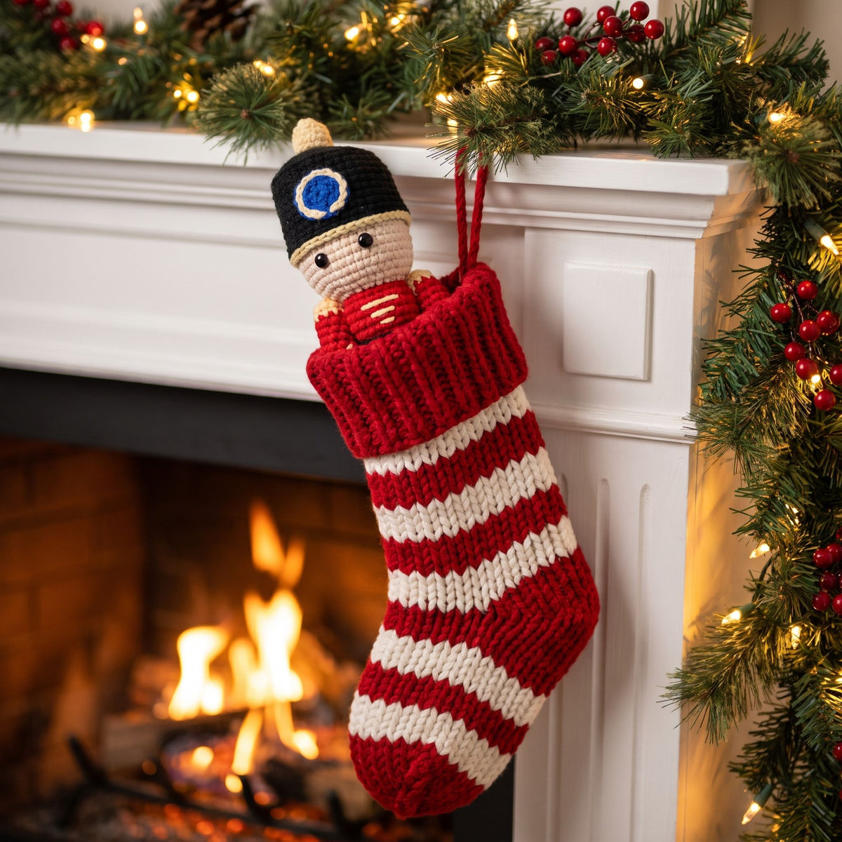 A hand-knit Christmas stocking hanging from a white fireplace mantel decorated with green pine garlands, red berries, and warm white string lights. The stocking features red and white horizontal stripes and is pulled out of a Handmade crochet full body nutcracker ornament by YarnYummies front view showing uniform detail, black knitted hat with a blue circular emblem and a cream-colored face peeking out.