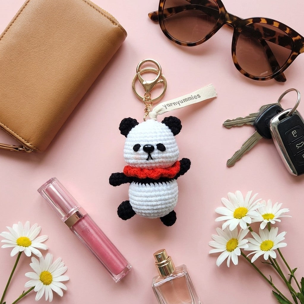 Panda keychain with a red bow tie on a pink surface with flowers, keys, and sunglasses.
