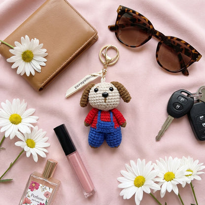 Dog-shaped keychain with red sweater and blue overalls, surrounded by daisies, wallet, sunglasses, keys, and perfume bottle on a pink background.