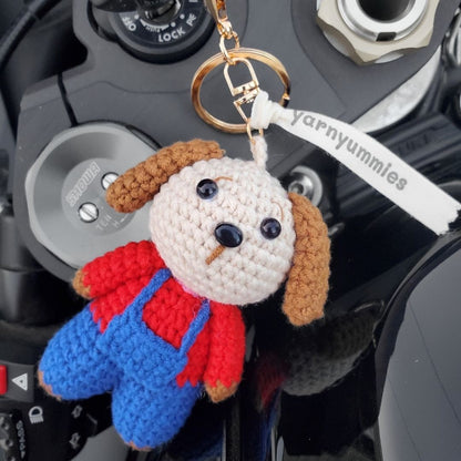 Crocheted dog keychain with red sweater and blue overalls on a car dashboard, featuring 'yarnyummies' brand.