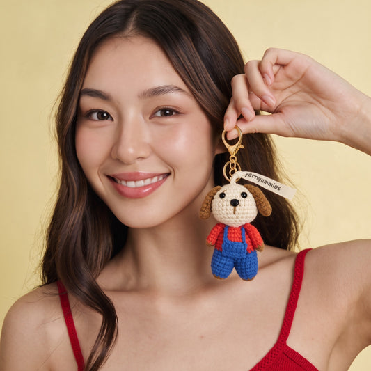 A smiling woman holds a crocheted dog keychain charm against a soft yellow background. The charm features a cream-colored head, brown floppy ears, a red top, and blue pants, with a gold key ring and a small tag that reads yarnyummies.