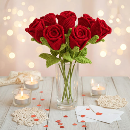 Bouquet of red knitted roses in a glass vase on a wooden table with candles and heart-shaped decorations.