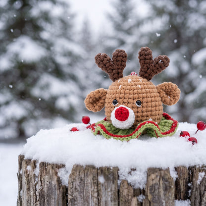 A Handmade crochet reindeer head ornament with rattle by YarnYummies front view with brown antlers, a tan face, a red nose, and a green and red ruffled collar, placed on a snowy log outdoors in the snow.