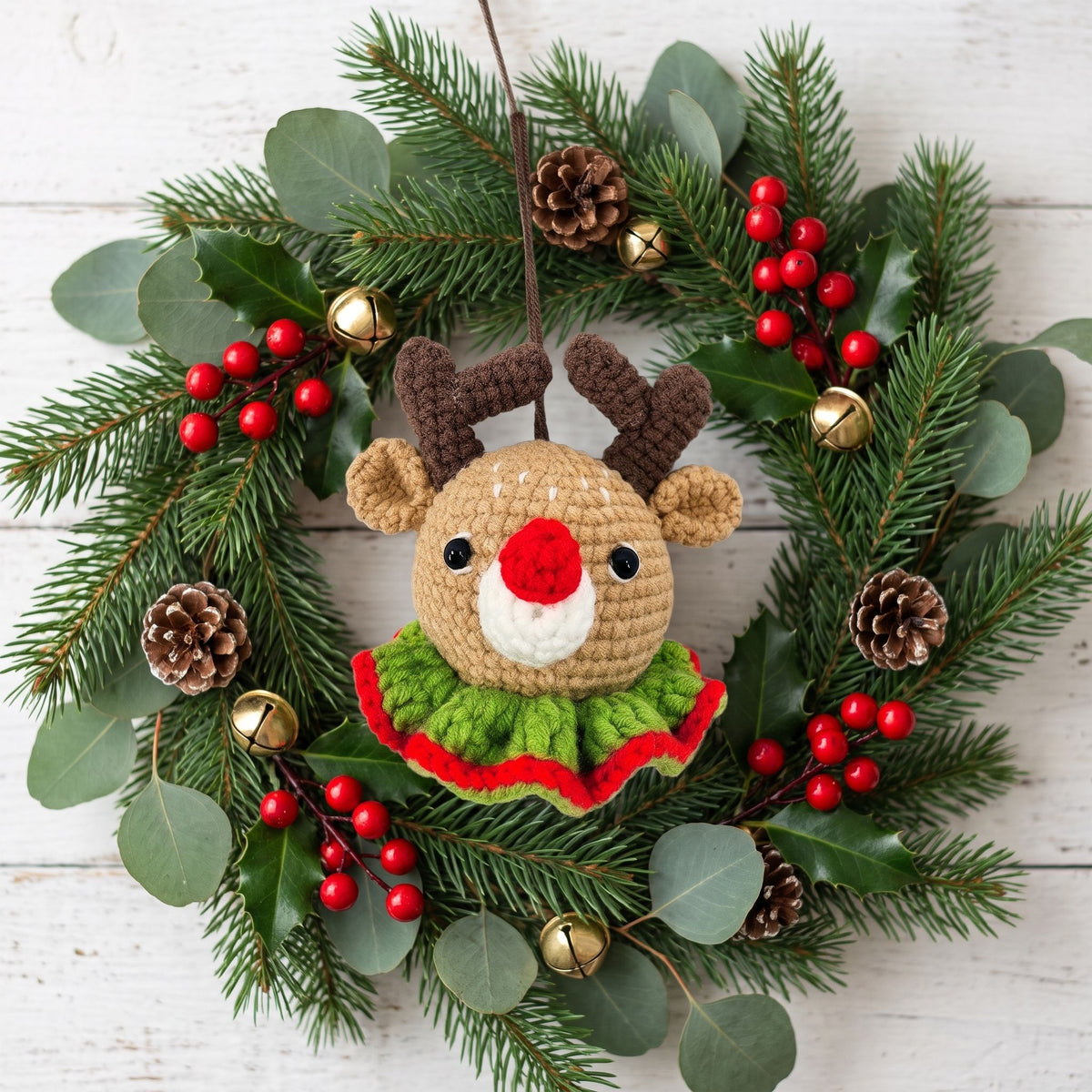 A crocheted Christmas wreath featuring a Handmade crochet reindeer head ornament by YarnYummies front view showing red nose and antlers in the center, surrounded by green pine branches, eucalyptus leaves, red berries, pinecones, and small gold bells on a white wood plank background.