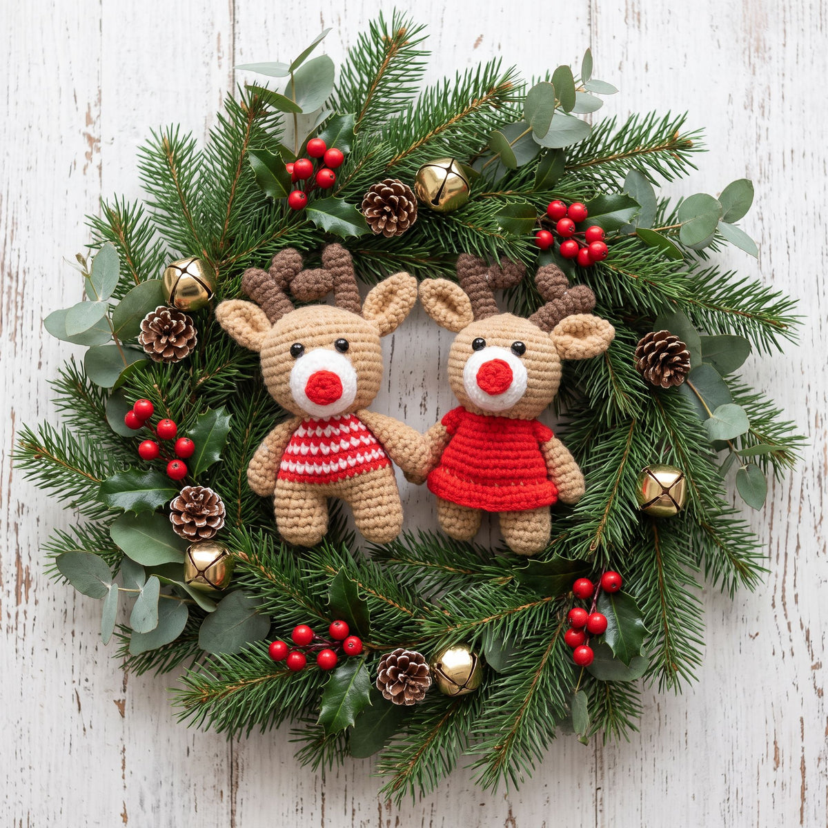 A festive Christmas wreath featuring Handmade crochet reindeer couple ornament set by YarnYummies front view showing both reindeer with coordinated outfits surrounded by green pine branches, eucalyptus leaves, red berries, pinecones, and gold bells on a whitewashed wooden background.