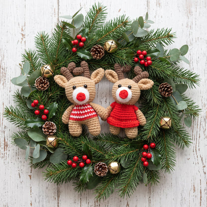 A festive Christmas wreath featuring Handmade crochet reindeer couple ornament set by YarnYummies front view showing both reindeer with coordinated outfits surrounded by green pine branches, eucalyptus leaves, red berries, pinecones, and gold bells on a whitewashed wooden background.