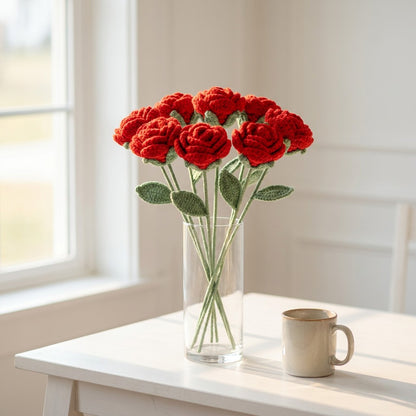 Vase with red crocheted roses on a white table next to a mug.