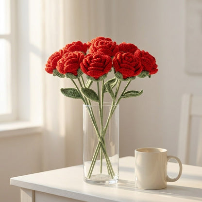 Vase with red crocheted roses on a table next to a white mug.