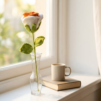 Crocheted rose flower in a clear vase on a windowsill with a book and mug.