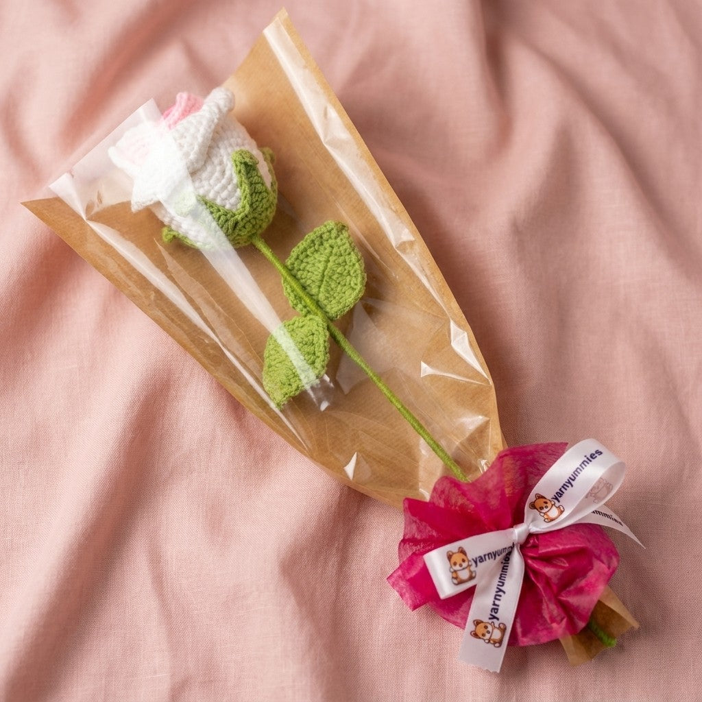 Crocheted white and pink rose with green leaves in clear packaging on a pink fabric background