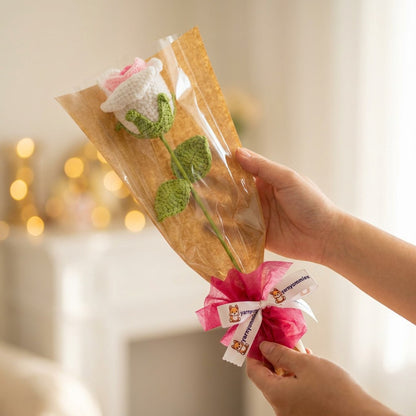 Hand holding a small bouquet of flower wrapped in clear plastic with a decorative bow against a blurred background.
