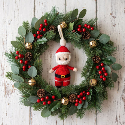 A festive Christmas wreath made of green pine branches with eucalyptus leaves, red berries, pinecones, and small gold bells, decorated with a handmade crocheted baby safe Santa Claus ornament by YarnYummies hanging in the center on a whitewashed wood background.