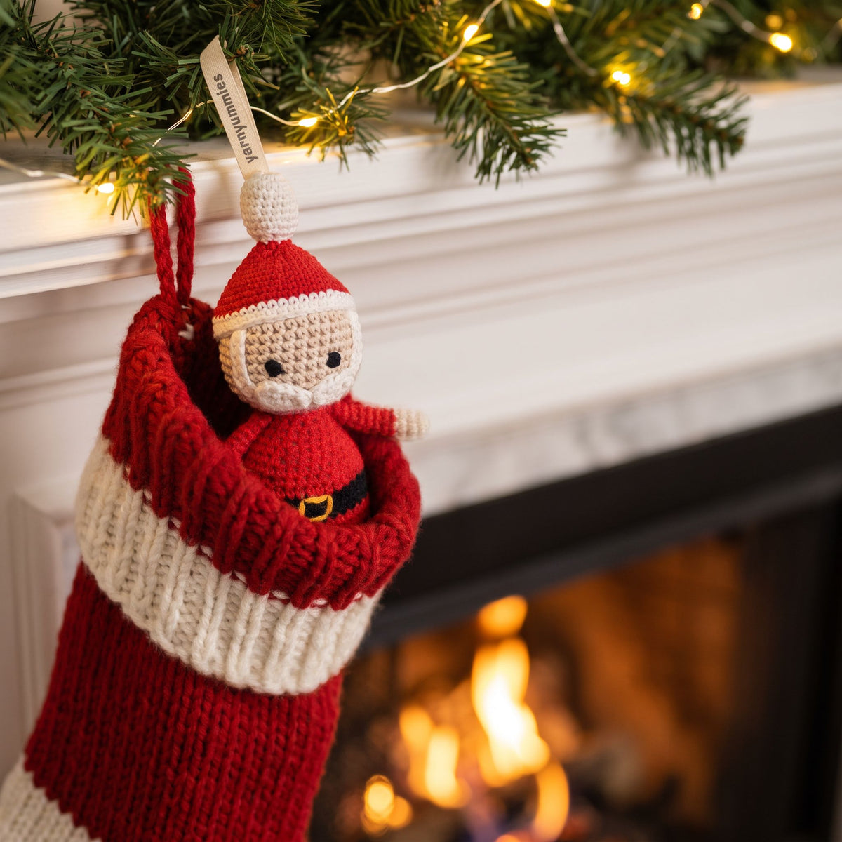 A red and cream knitted Christmas stocking hangs from a white fireplace mantel decorated with green pine branches and warm string lights. A handmade crocheted baby safe Santa ornament by YarnYummies peeks out of the stocking, with a glowing fireplace in the background. 