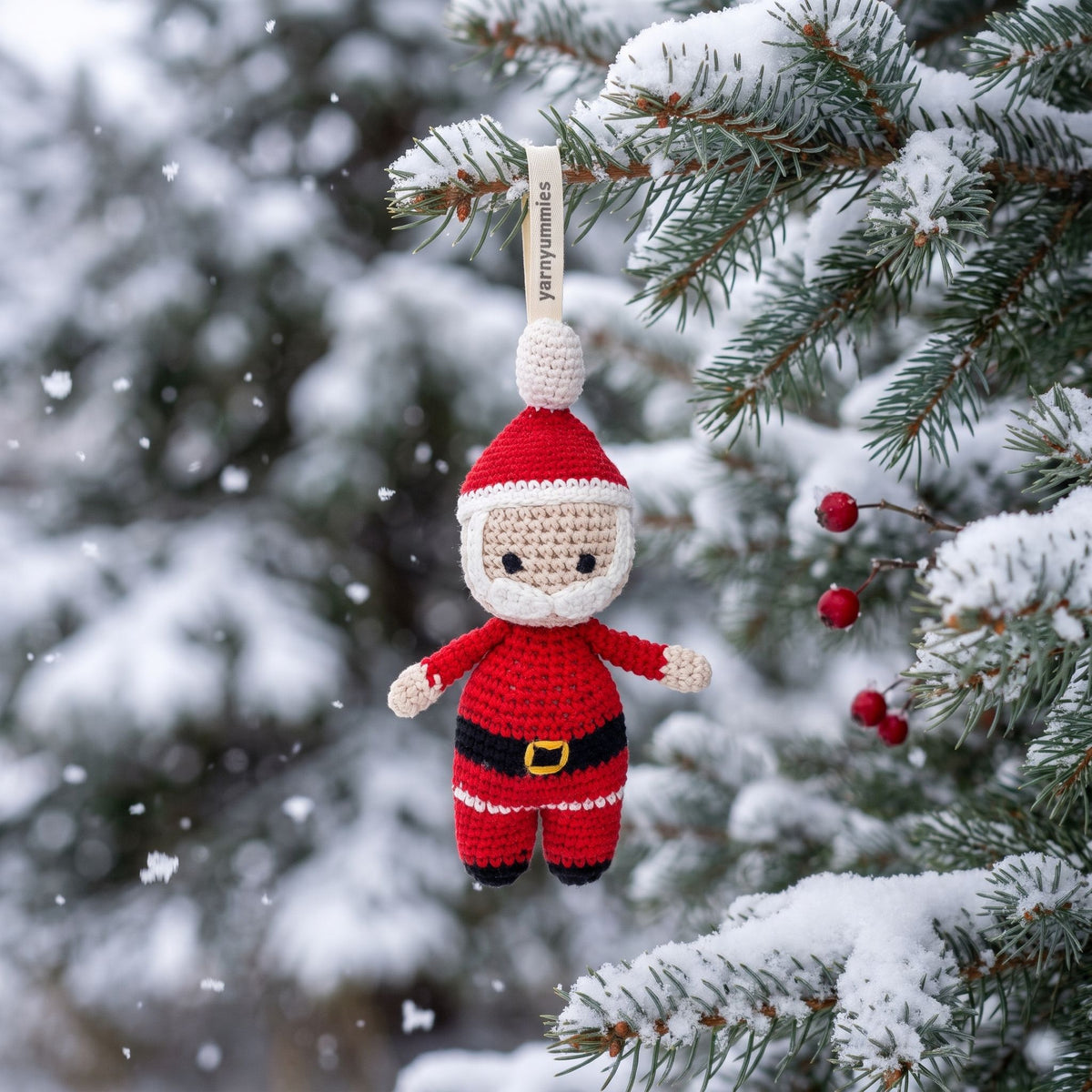 A handmade crocheted baby safe Santa Claus hanging ornament by YarnYummies is suspended from a snow-covered evergreen tree branch, with falling snow and blurred pine trees in the background.