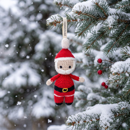 A handmade crocheted baby safe Santa Claus hanging ornament by YarnYummies is suspended from a snow-covered evergreen tree branch, with falling snow and blurred pine trees in the background.
