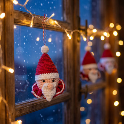 A Handmade crochet Santa head ornament with rattle by YarnYummies front view hangs from a wooden window frame decorated with warm string lights and blurred festive lights in the background, with snow visible outside.