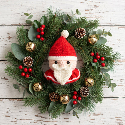 A festive Christmas wreath featuring a central Handmade crochet Santa head ornament by YarnYummies front view, surrounded by green pine branches, eucalyptus leaves, red berries, pinecones, and small gold bells, arranged on a distressed white wooden background.