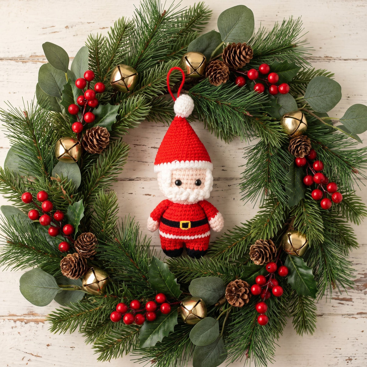 A festive Christmas wreath made of green pine branches, decorated with red berries, pinecones, gold bells, and handmade crochet full body Santa ornament by YarnYummies in the center, set against a whitewashed wooden background.