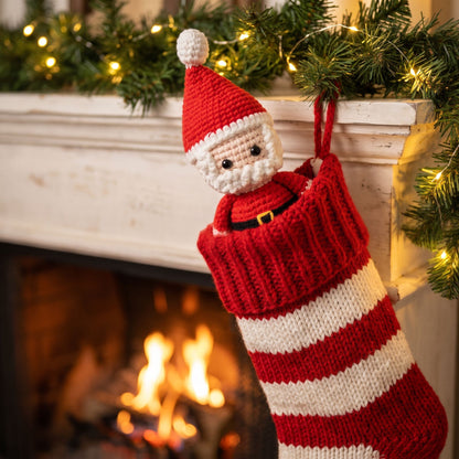 A knitted Christmas stocking with a Handmade crochet full body Santa ornament by YarnYummies hanging from a white fireplace mantel, with a warm fire burning below and green garland with string lights above.