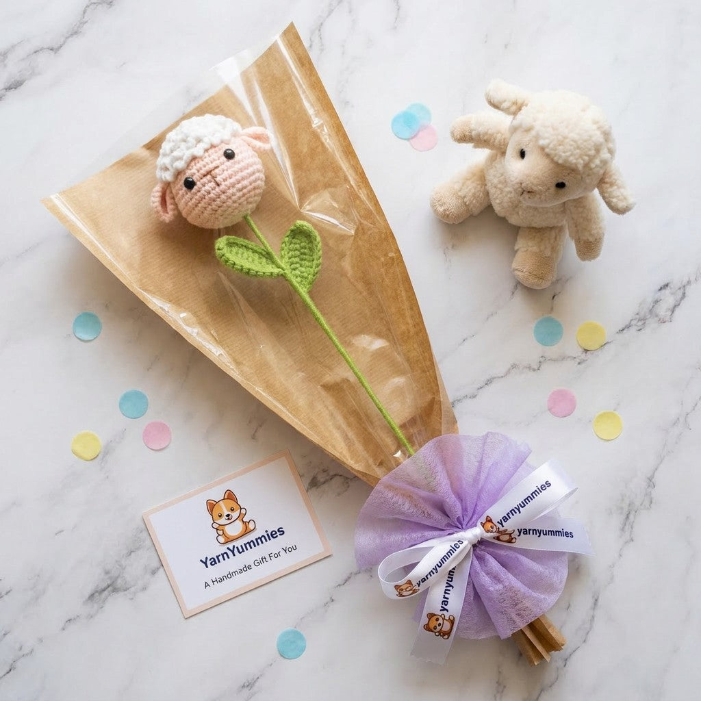 Yarn Yummies product with a small sheep toy, crocheted flower shaped like a sheep head, and card on a marble surface.