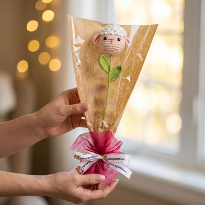 Hand holding a bouquet of knitted flowers shaped like a head of a sheep in a clear bag, against a blurred indoor background.