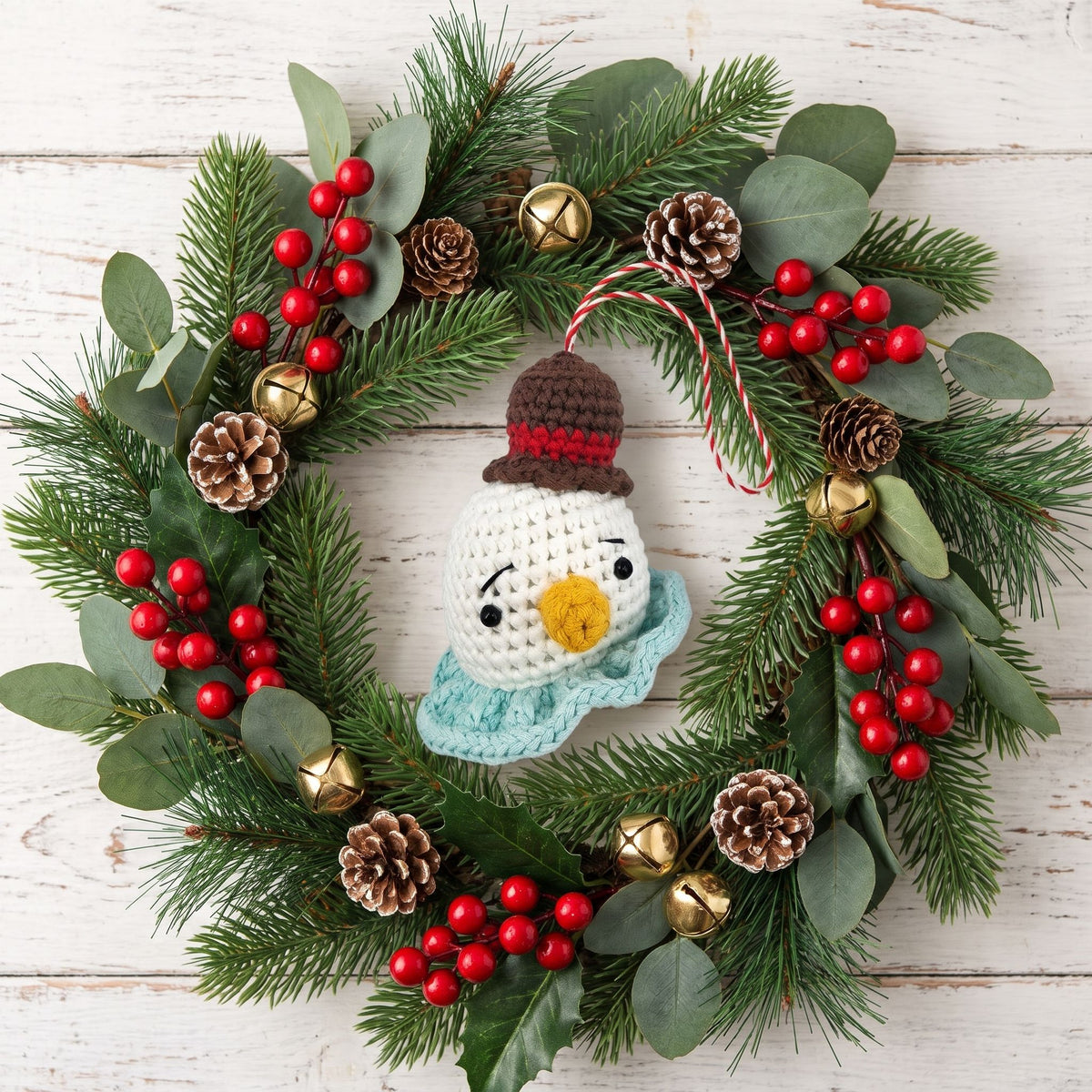 A festive Christmas wreath made of green pine branches, eucalyptus leaves, and red berries, decorated with small gold bells, pinecones, and a Handmade crochet snowman head ornament with rattle by YarnYummies in the center, set against a white wood plank background.