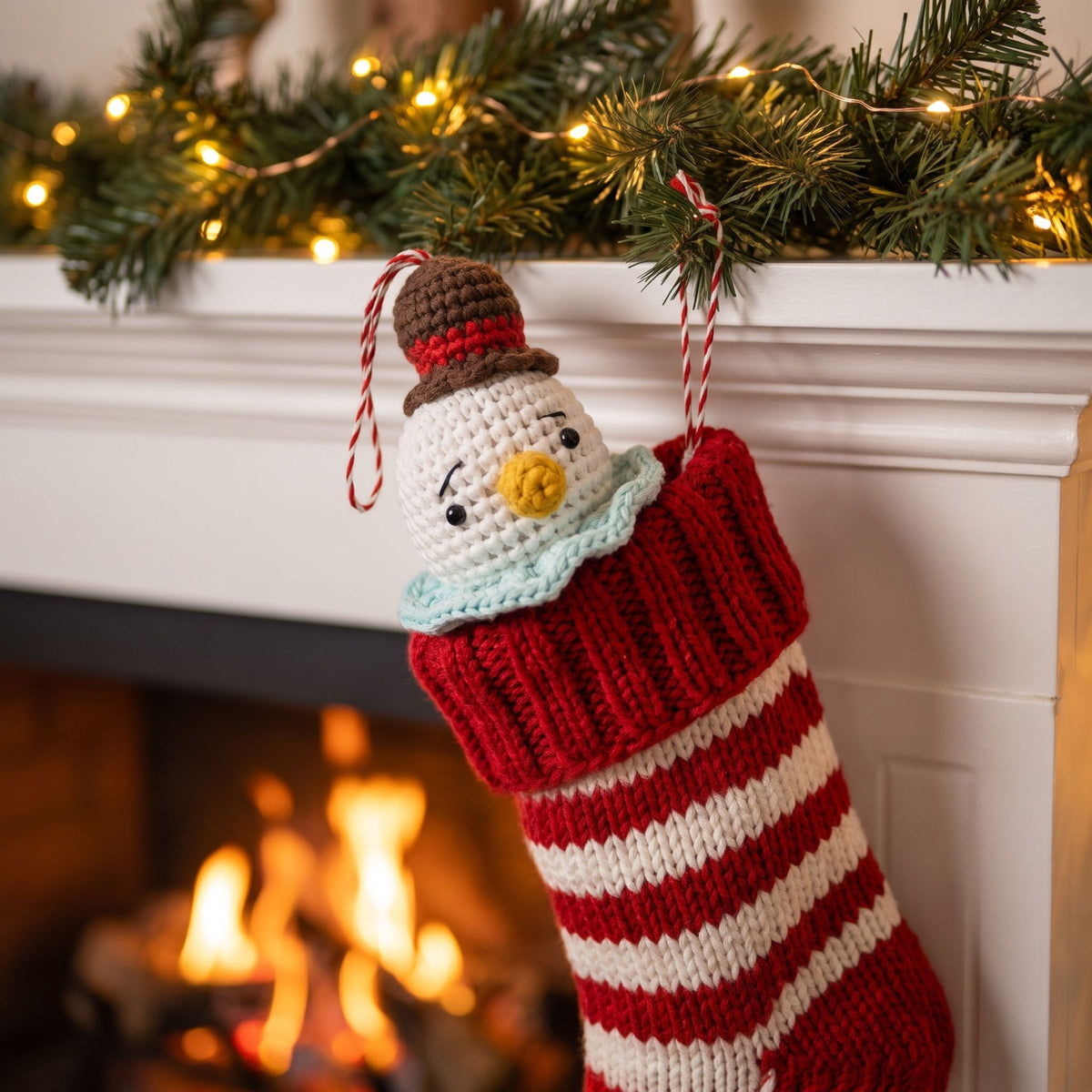 A knitted Christmas stocking with a Handmade crochet snowman head ornament with rattle by YarnYummies peeking out, hangs from a white fireplace mantel decorated with green garland and warm string lights, with a fire burning below.