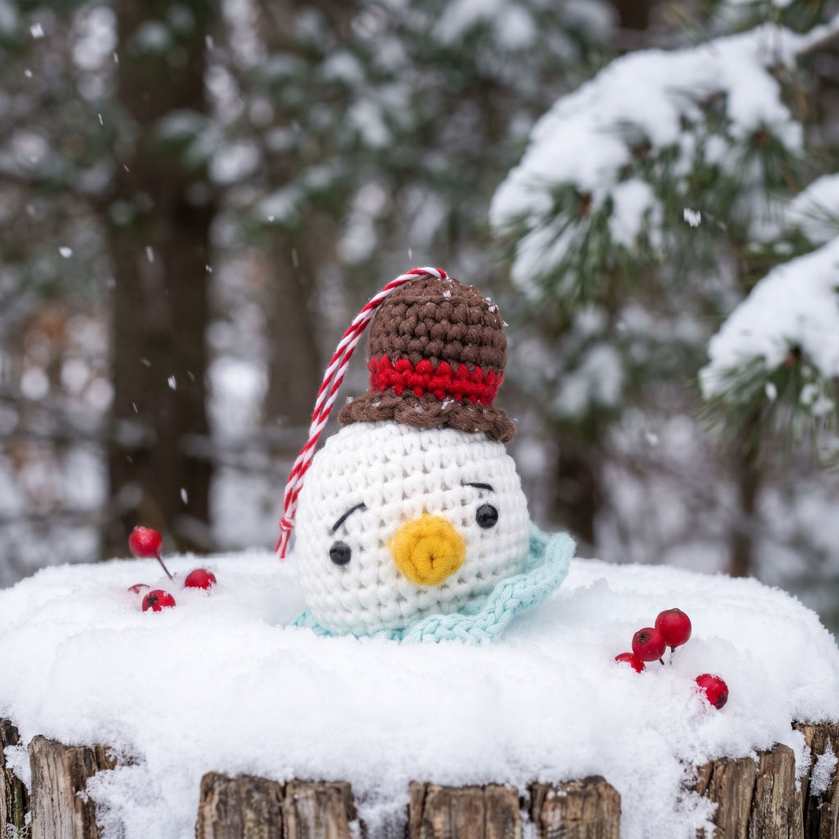 A Handmade crochet snowman head ornament with rattle by YarnYummies wearing a brown and red hat with a red-and-white striped candy cane string, a light blue scarf, and a yellow nose, sitting outdoors on a snowy log with snow-covered trees in the background.