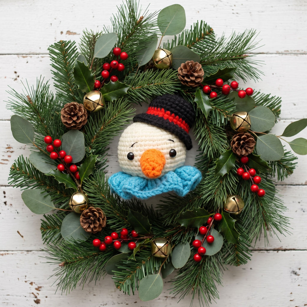 A festive Handmade crochet snowman head ornament by YarnYummies front view showing carrot nose and festive hat centered on a green pine wreath decorated with red berries, pinecones, eucalyptus leaves, and small gold bells, set against a white wood plank background.