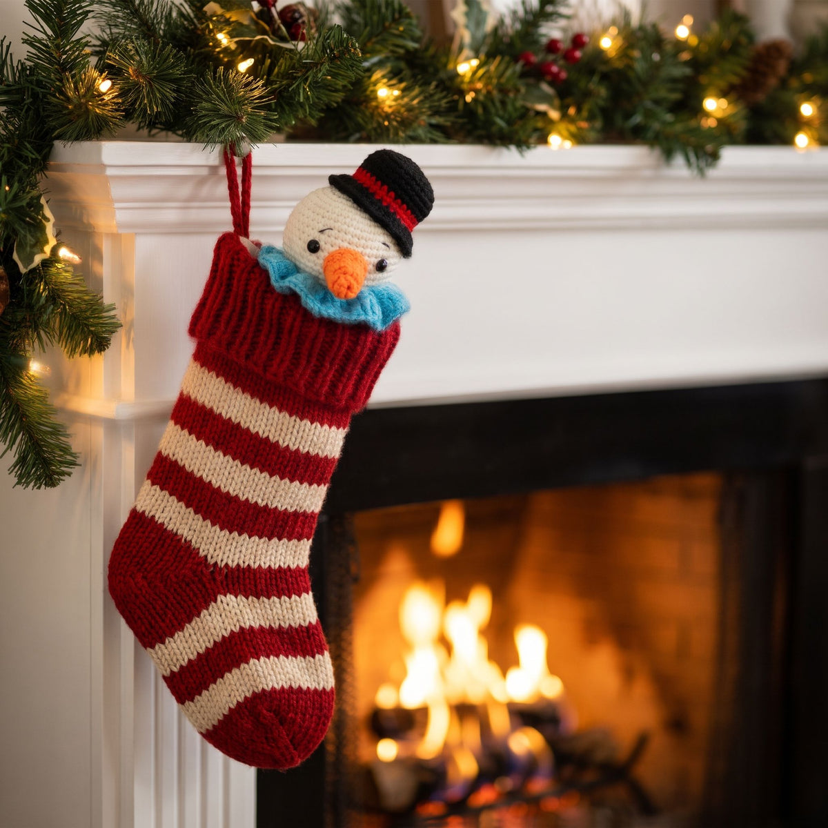 A red and cream striped knit Christmas stocking with a Handmade crochet snowman head ornament by YarnYummies front view showing carrot nose and festive hat peeking out, hanging from a white fireplace mantel decorated with green garland and warm string lights, with a burning fireplace in the background.