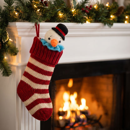 A red and cream striped knit Christmas stocking with a Handmade crochet snowman head ornament by YarnYummies front view showing carrot nose and festive hat peeking out, hanging from a white fireplace mantel decorated with green garland and warm string lights, with a burning fireplace in the background.