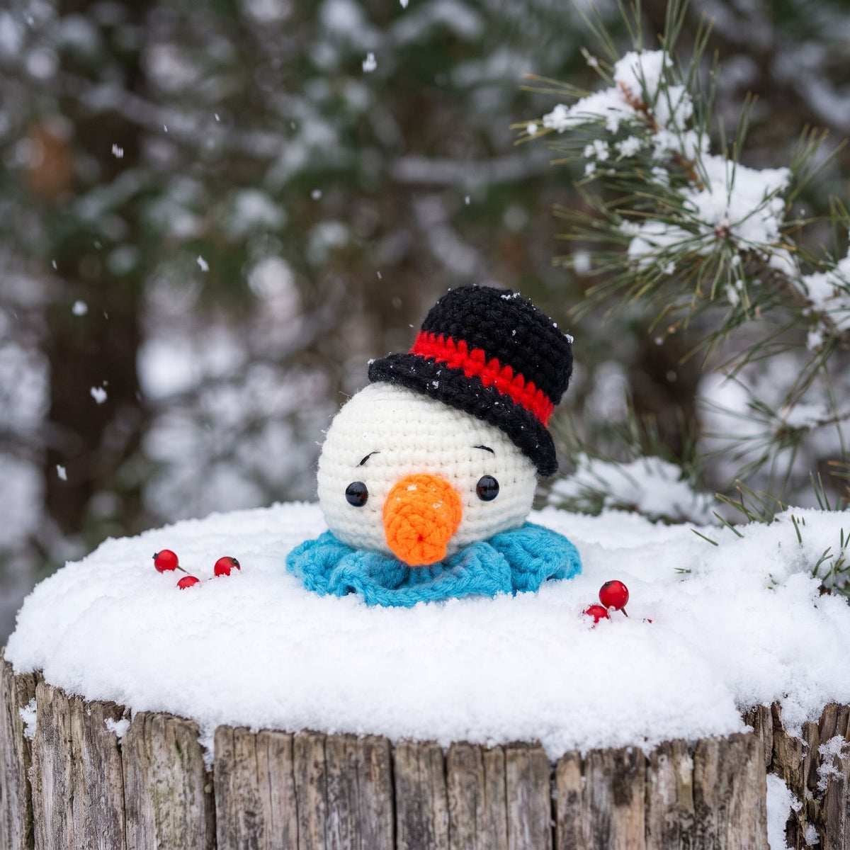 A Handmade crochet snowman head ornament by YarnYummies front view showing carrot nose and festive hat sitting on a snowy log outdoors in the winter, wearing a black hat with a red band, a blue scarf, and an orange nose. Snow is falling in the background with pine branches visible.