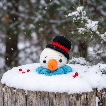 A Handmade crochet snowman head ornament by YarnYummies front view showing carrot nose and festive hat sitting on a snowy log outdoors in the winter, wearing a black hat with a red band, a blue scarf, and an orange nose. Snow is falling in the background with pine branches visible.