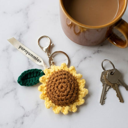 Crocheted sunflower keychain with a leaf, keys, and a mug on a marble surface.